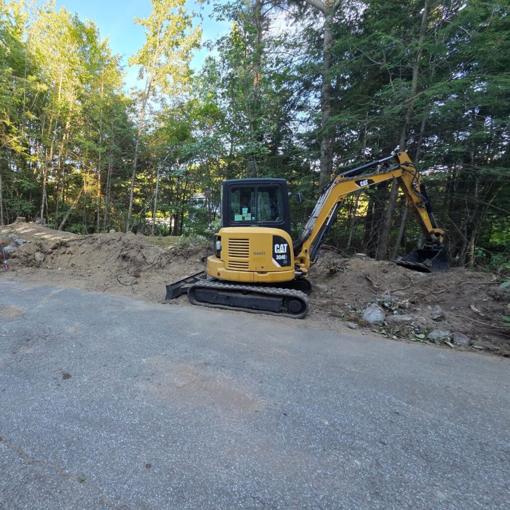 Utility trenching in Central Massachusetts with a compact excavator cutting a service line trench along a driveway edge